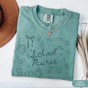 May include: Light green Comfort Colors t-shirt with the words "School Nurse" and school-themed doodles. The shirt has a crew neck and short sleeves. A gold necklace with a pendant is draped across the shirt.