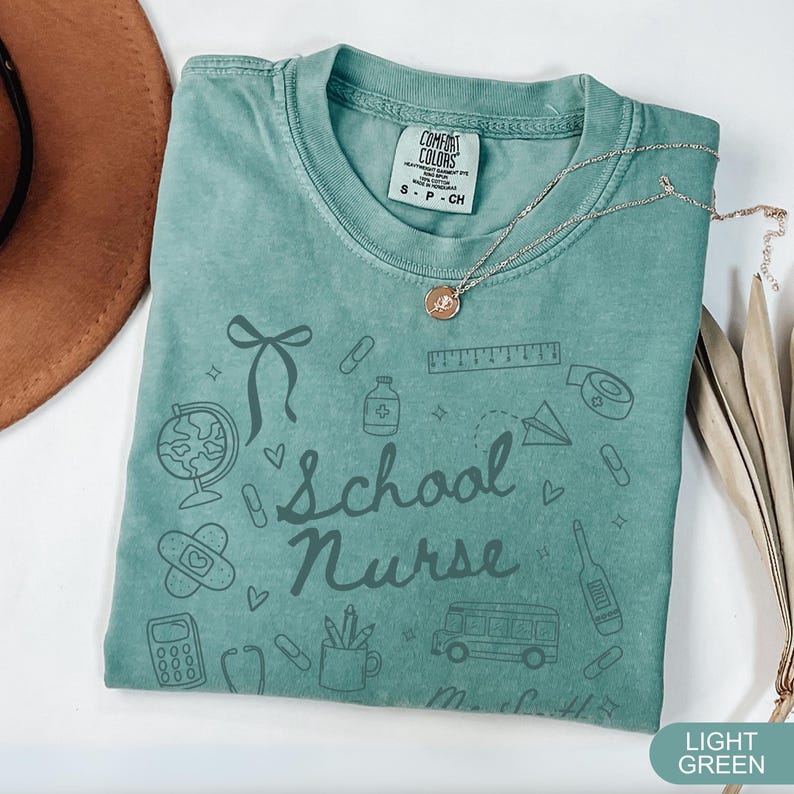 May include: Light green Comfort Colors t-shirt with the words "School Nurse" and school-themed doodles. The shirt has a crew neck and short sleeves. A gold necklace with a pendant is draped across the shirt.