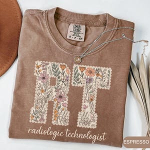 May include: Espresso-colored t-shirt with the letters "RT" filled with floral designs. The words "radiologic technologist" are printed below the letters. A silver necklace with a pendant rests on the shirt.