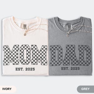 May include: Two t-shirts, ivory and grey, with the words "MOM" and "DAD" in a checkered pattern. The text "EST. 2025" is below each word. Each shirt has a silver necklace. The ivory shirt is on the left, and the grey shirt is on the right.