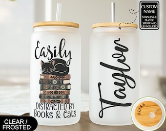 May include: Two frosted glass tumblers with bamboo lids and straws. One tumbler has a black cat sleeping on a stack of books with the text "Easily Distracted by Books & Cats". The other tumbler has the name "Taylor" written on it. Both tumblers have a clear plastic straw and a bamboo lid.