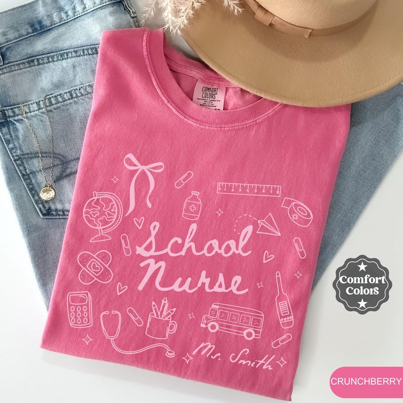 May include: A pink t-shirt with the words "School Nurse" and related graphics, including a globe, band-aid, and bus. The shirt is paired with blue jeans and a tan hat. The shirt is labeled "Comfort Colors" and the color is "Crunchberry".