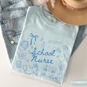May include: Light blue t-shirt with a school nurse design in blue. The design includes a globe, band-aid, calculator, stethoscope, bus, and the words "School Nurse" and "Mr. Smith". A tan hat and blue jeans are in the background.