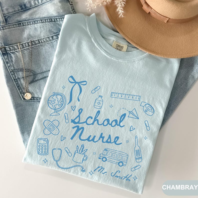 May include: Light blue t-shirt with a school nurse design in blue. The design includes a globe, band-aid, calculator, stethoscope, bus, and the words "School Nurse" and "Mr. Smith". A tan hat and blue jeans are in the background.