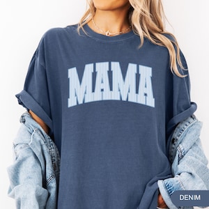 以下が含まれることがあります： デニムブルーのTシャツ。大きなライトブルーの縦縞模様の文字で「MAMA」と書かれています。 ゆったりとしたフィット感で、袖はロールアップされています。 右下には「DENIM」の文字がプリントされています。