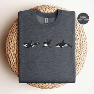 May include: A dark heather grey t-shirt featuring a graphic of three black and white orcas swimming in a row. The text "DARK HEATHER" is in the top right corner of the image.