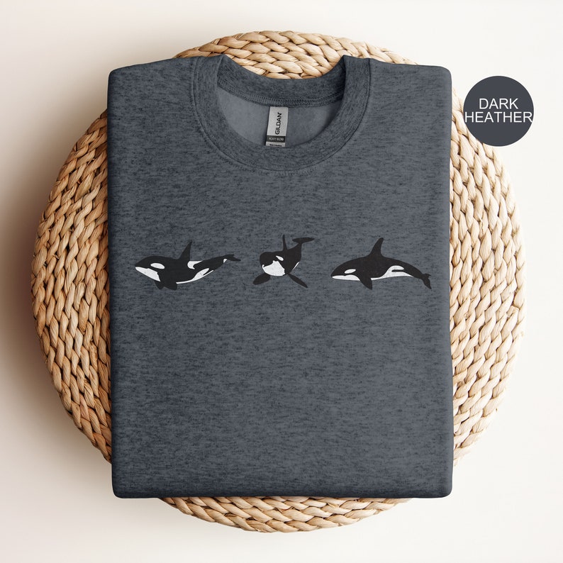 May include: A dark heather grey t-shirt featuring a graphic of three black and white orcas swimming in a row. The text "DARK HEATHER" is in the top right corner of the image.