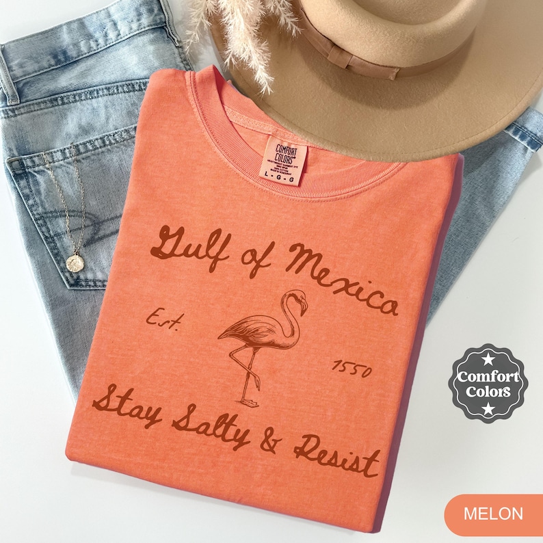 Peut inclure: Un t-shirt orange corail avec un graphique vintage d'un flamant rose et le texte "Gulf of Mexico Est. 1550 Stay Salty & Resist". Le t-shirt a un aspect vieilli et est fabriqu&eacute; par Comfort Colors.