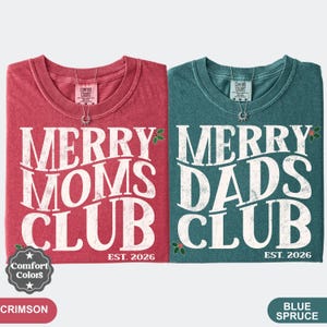 Könnte beinhalten: Zwei kurzärmelige T-Shirts, eines in Karmesinrot und eines in Blau-Fichte, mit weißem, abgenutztem Text, der MERRY MOMS CLUB EST. 2026 und MERRY DADS CLUB EST. 2026 lautet. Beide Shirts zeigen kleine grüne Stechpalmenblätter und rote Beeren.