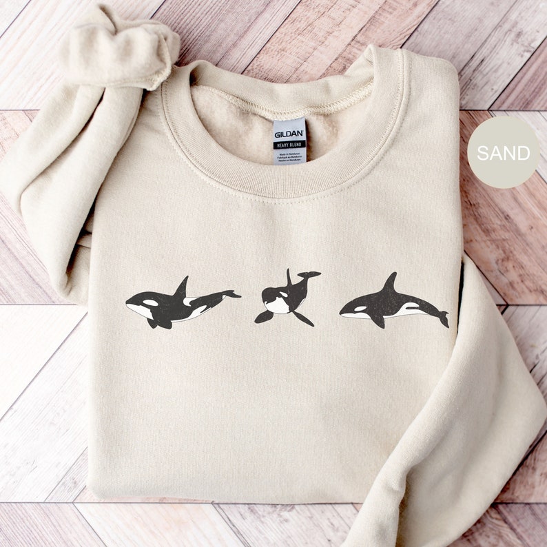 May include: A sand-coloured sweatshirt with a graphic of three black and white orcas swimming in a row. The sweatshirt has a Gildan tag on the bottom.