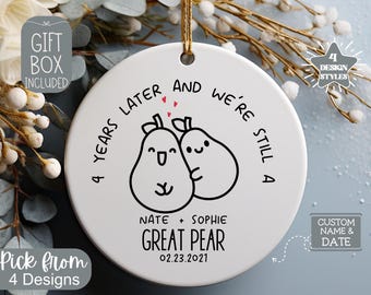 May include: White ceramic ornament with a black and white illustration of two pears with faces. The text reads "4 years later and we're still a great pear" with the names "Nate + Sophie" and the date "02.23.2021".