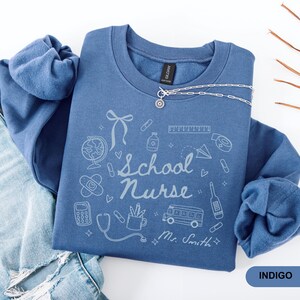 May include: Indigo blue sweatshirt with a school nurse design. The design includes a globe, band-aid, calculator, stethoscope, bus, and the words "School Nurse" and "Mr. Smith". A silver necklace is draped across the sweatshirt. The word "INDIGO" is in a blue rectangle.