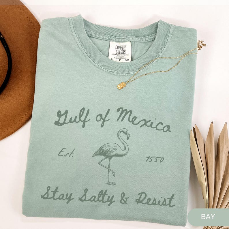 Peut inclure: Un t-shirt vert clair avec un graphique vintage d'un flamant rose et le texte "Gulf of Mexico Est. 1950 Stay Salty & Resist".