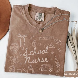 May include: Espresso-colored t-shirt with the words "School Nurse" and related graphics. The shirt features a crew neck and short sleeves. A silver necklace with a pendant is draped across the shirt.