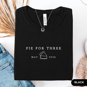 Może przedstawiać: Czarny t-shirt z napisem "PIE FOR THREE" nad grafiką przedstawiającą kawałek ciasta. Poniżej grafiki znajduje się napis "MAY 2026". Srebrny naszyjnik z zawieszką w kształcie słońca leży na koszulce.
