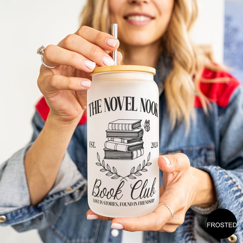 K&ouml;nnte beinhalten: Ein gefrosteter Glasbecher mit Bambusdeckel und Strohhalm. Der Becher hat ein schwarz-wei&szlig;es Design mit dem Text "The Novel Nook Book Club Est. 2024 Lost in stories, found in friendship".
