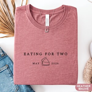 Może przedstawiać: Koszulka w kolorze wrzosowym z napisem "EATING FOR TWO" i datą "MAJ 2026" z grafiką ciasta. Koszulka jest złożona i ma okrągły dekolt. Widoczny jest naszyjnik z zawieszką w kształcie słońca.