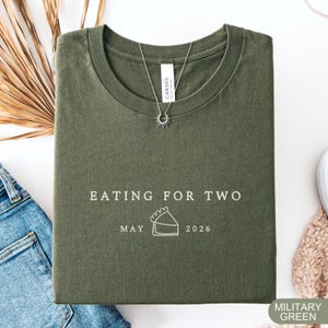 Może przedstawiać: Wojskowa zielona koszulka z napisem "EATING FOR TWO" i datą "MAJ 2026" pod grafiką ciasta. Srebrny naszyjnik z zawieszką w kształcie słońca leży na koszulce. Koszulka jest złożona na białej powierzchni.