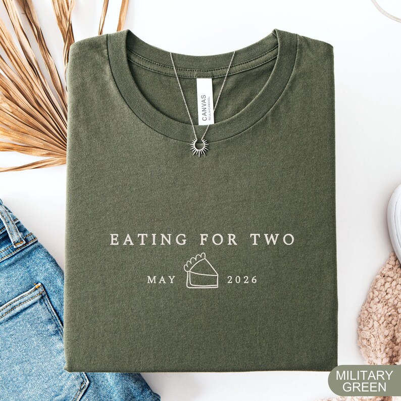 Może przedstawiać: Wojskowa zielona koszulka z napisem "EATING FOR TWO" i datą "MAJ 2026" pod grafiką ciasta. Srebrny naszyjnik z zawieszką w kształcie słońca leży na koszulce. Koszulka jest złożona na białej powierzchni.