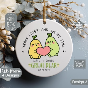 May include: A white ceramic ornament with a gold string for hanging. The ornament features two cartoon pears holding a pink heart, with text that reads "4 years later and we're still a great pear." Includes the names Nate and Sophie, and the date 02.23.2021.