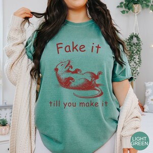May include: A teal blue t-shirt with a red graphic of a possum and the text "Fake it till you make it".