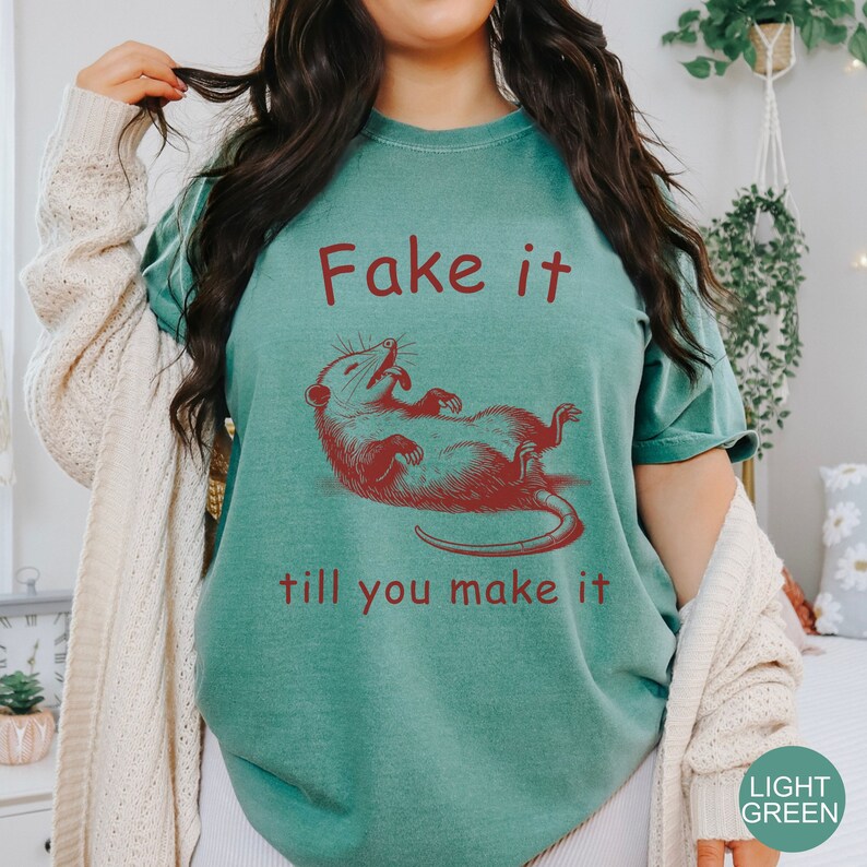 May include: A teal blue t-shirt with a red graphic of a possum and the text "Fake it till you make it".
