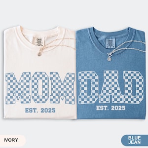 May include: Two t-shirts, one ivory and one blue jean, with the words "MOM" and "DAD" in a checkered pattern. The text "EST. 2025" is below each word. Each shirt has a silver necklace. The bottom of the image has the color names.