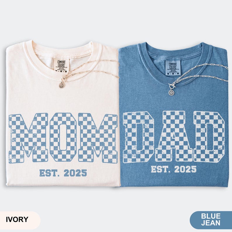 May include: Two t-shirts, one ivory and one blue jean, with the words "MOM" and "DAD" in a checkered pattern. The text "EST. 2025" is below each word. Each shirt has a silver necklace. The bottom of the image has the color names.