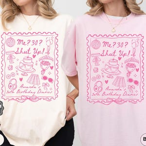 May include: Two t-shirts with a pink graphic design. The graphic features a pink stamp with the text "Me? 30? Shut Up!" and illustrations of a cake, a gift, and a pair of sunglasses. The text "Amanda's 30th Birthday Diaries" is below the stamp. One t-shirt is ivory and the other is pink.