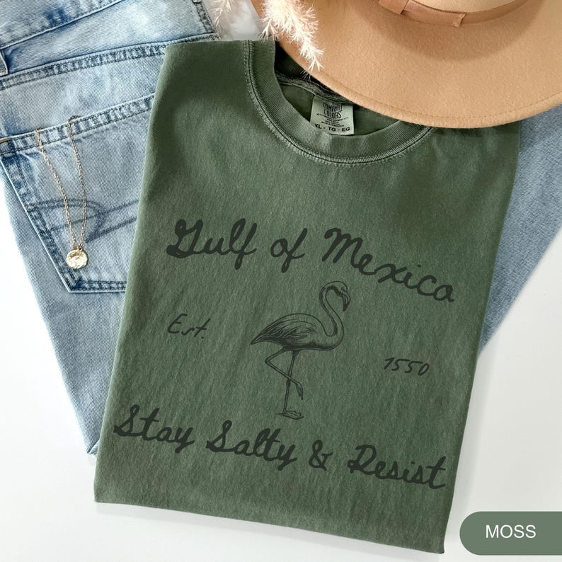 Peut inclure: Un t-shirt vert avec le texte "Gulf of Mexico Est. 1550 Stay Salty & Resist" et une illustration de flamant rose noir. Le t-shirt est sur un fond en jean bleu clair.