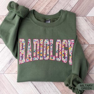 May include: A green sweatshirt with the word "RADIOLOGY" printed in white letters outlined in pink. The letters are filled with a floral pattern.