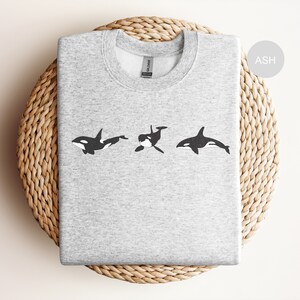 May include: A grey sweatshirt with a black and white graphic of three orcas swimming in a row. The text "ASH" is in the top right corner of the image.