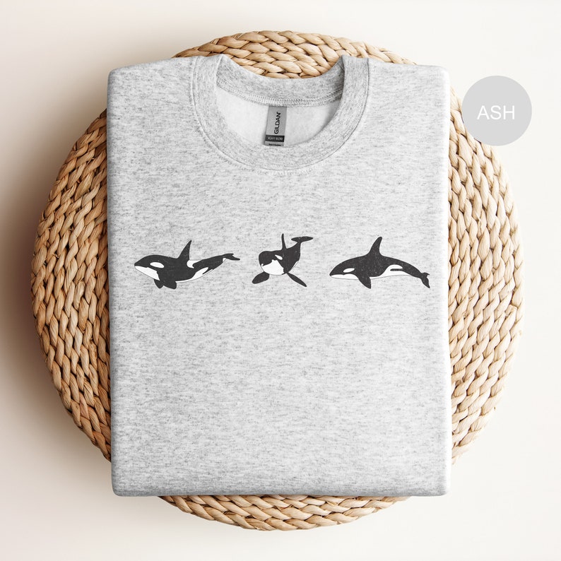 May include: A grey sweatshirt with a black and white graphic of three orcas swimming in a row. The text "ASH" is in the top right corner of the image.