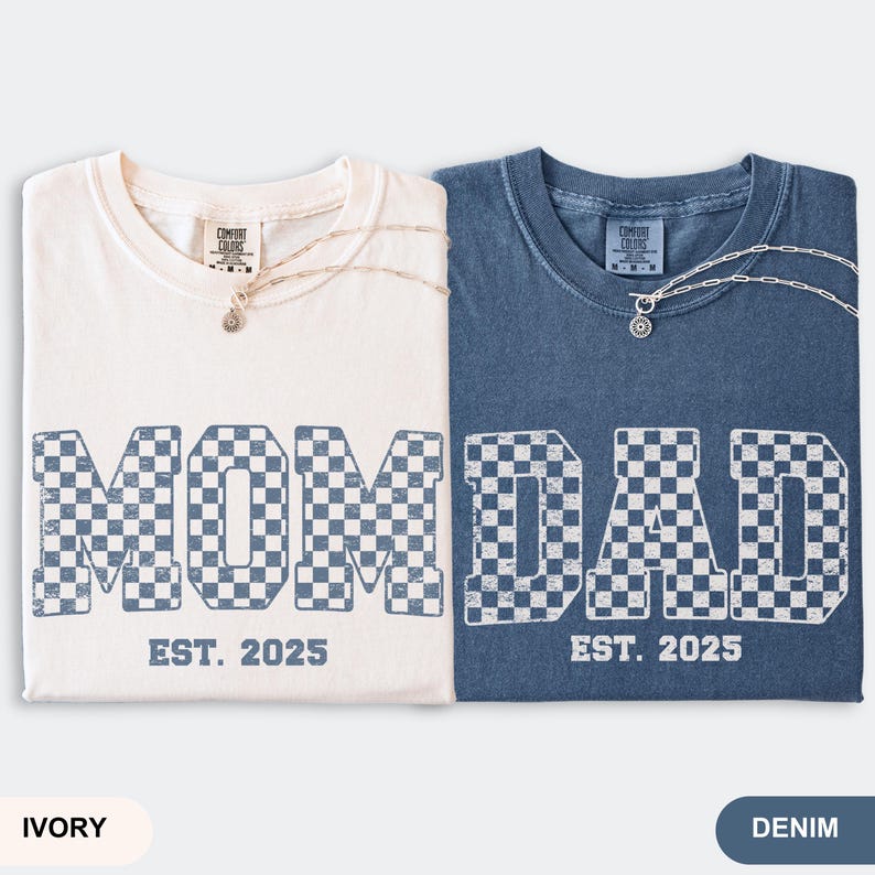 May include: Two t-shirts, ivory and denim, with the words "MOM" and "DAD" in a checkered pattern. The text "EST. 2025" is below the words. Each shirt has a silver necklace with a pendant. The ivory shirt is on the left, and the denim shirt is on the right.