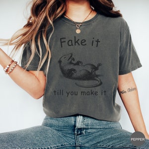 May include: A gray t-shirt with a black and white illustration of a possum and the text "Fake it till you make it".