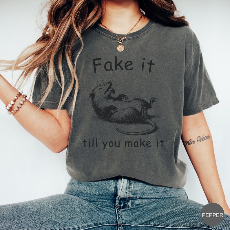 May include: A gray t-shirt with a black and white illustration of a possum and the text "Fake it till you make it".