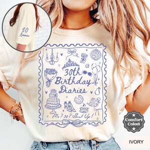 May include: A white t-shirt with a blue graphic design featuring a stamp with the text "30th Birthday Diaries" and the words "Me? 30? Shut Up!" The graphic also includes various illustrations such as a cake, a crown, a teacup, and a pair of shoes. The shirt has the words "Comfort Colors" and "IVORY" printed on the bottom right corner.