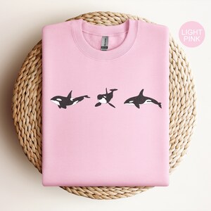 May include: A light pink sweatshirt featuring a design of three black and white orcas swimming in a row.