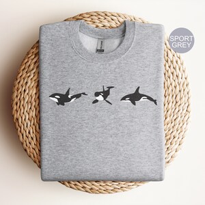 May include: A grey sweatshirt featuring a black and white graphic of three orcas swimming in a row. The text "SPORT GREY" is in the top right corner of the image.
