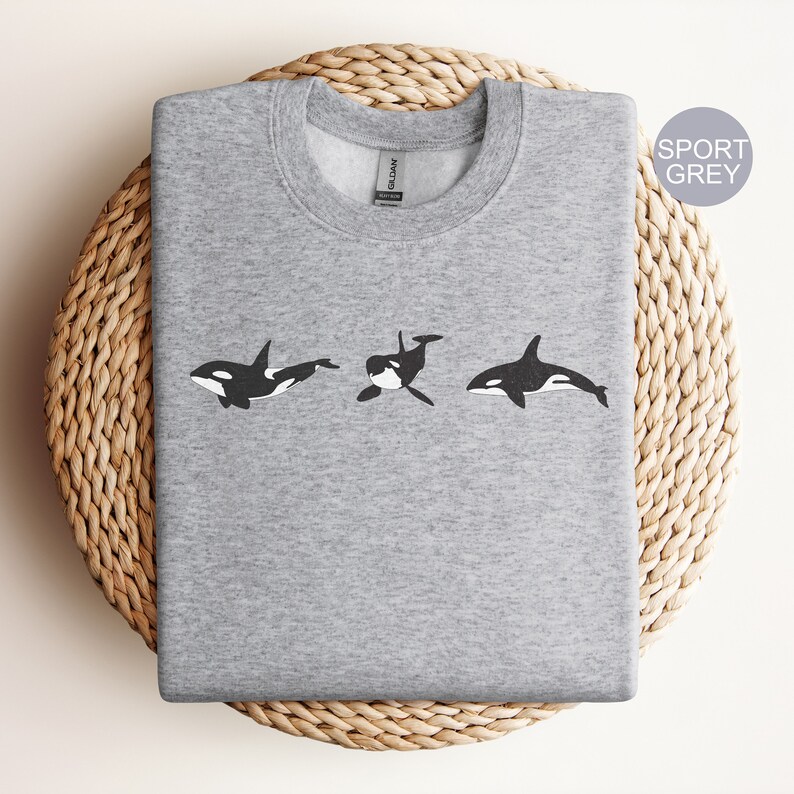 May include: A grey sweatshirt featuring a black and white graphic of three orcas swimming in a row. The text "SPORT GREY" is in the top right corner of the image.