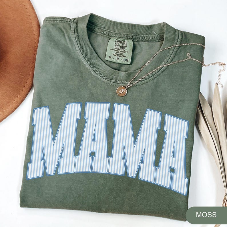 以下が含まれることがあります： モスグリーンのComfort Colors Tシャツ。大きな白い文字で「MAMA」と書かれ、水色の縦縞模様が入っています。金のネックレスとペンダントがシャツの上にかけられています。シャツは白い面に折りたたまれています。