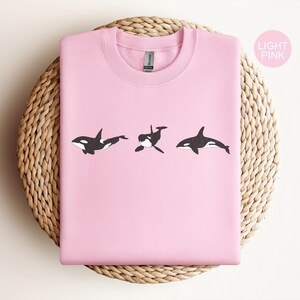 May include: A light pink sweatshirt with a graphic of three black and white orcas swimming in a row.