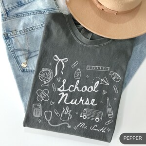 May include: A dark gray t-shirt with the words "School Nurse" and various school and medical-themed illustrations in white. The shirt is folded on top of blue jeans, with a tan hat and a gold necklace visible.
