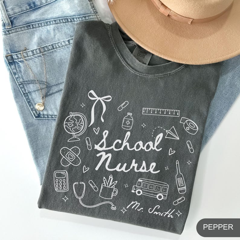 May include: A dark gray t-shirt with the words "School Nurse" and various school and medical-themed illustrations in white. The shirt is folded on top of blue jeans, with a tan hat and a gold necklace visible.