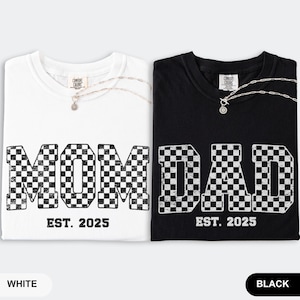 May include: Two t-shirts, one white and one black, with the words "MOM" and "DAD" in a checkered pattern. The text "EST. 2025" is below each word. Each shirt has a silver necklace. The white shirt is labeled "WHITE" and the black shirt is labeled "BLACK".