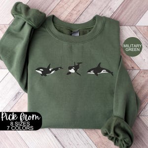 May include: A green sweatshirt with a graphic of three orcas in a row. The text "Pick from 8 sizes 7 colors" and "Military Green" are on the sweatshirt.
