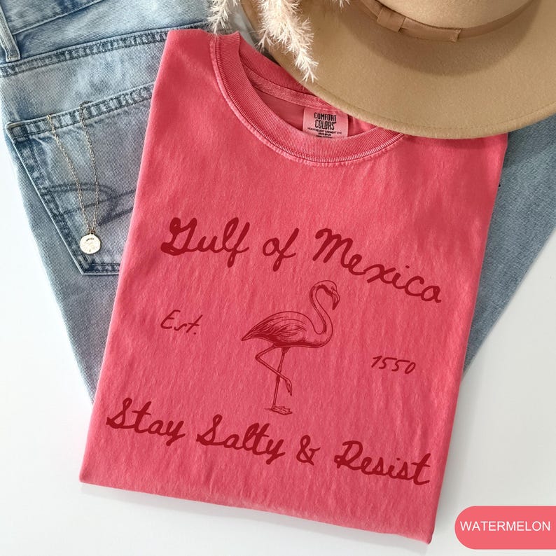 Peut inclure: Un t-shirt rouge avec un graphique de flamant rose et le texte "Gulf of Mexico Est. 1550 Stay Salty & Resist" en lettres rouges. Le t-shirt est pli&eacute; et pos&eacute; sur un jean bleu.