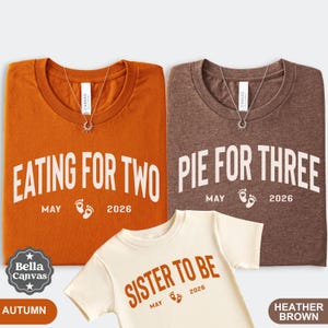 May include: Three t-shirts in autumn orange, heather brown, and cream. The orange shirt says "EATING FOR TWO" with "MAY 2026" below. The brown shirt says "PIE FOR THREE" with "MAY 2026" below. The cream shirt says "SISTER TO BE" with "MAY 2026" below.