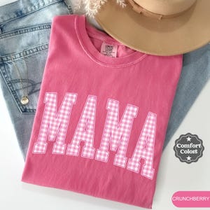 Könnte beinhalten: Ein pinkfarbenes T-Shirt mit dem Wort "MAMA" in weiß-rosa Vichy-Buchstaben. Das Shirt wird mit einer Jeans, einer Halskette und einem beigen Hut präsentiert. Das Shirt ist mit "Comfort Colors" gekennzeichnet und die Farbe ist "CRUNCHBERRY".