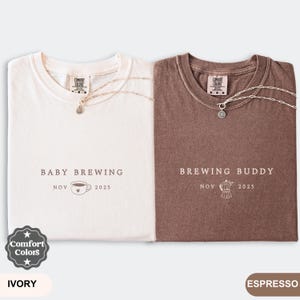 May include: Two Comfort Colors t-shirts, ivory and espresso, with matching silver necklaces. The ivory shirt reads "Baby Brewing" with a coffee cup graphic and "Nov 2025." The espresso shirt says "Brewing Buddy" with a coffee maker graphic and "Nov 2025."